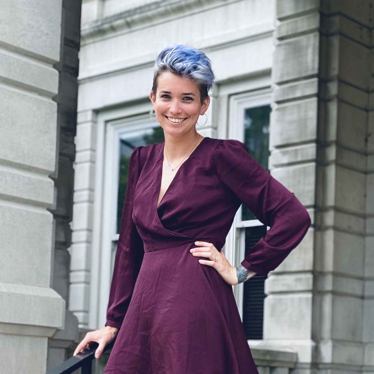 Agent Liv Ivanov standing in front of a grey building. She has short, blue hair and is wearing an eggplant-purple dress. She is smiling and has one hand on her hip.